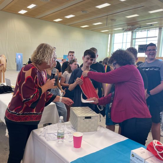 Eine Gruppe von Menschen ist um einen Tisch versammelt, wobei zwei Frauen einem jungen Jungen eine rote Tasche präsentieren. Eine Frau hält ein Mikrofon und spricht. Der Tisch hat eine weiße Tischdecke, einen Becher, eine Flasche und Kisten. Hinter ihnen stehen weitere Menschen, einer mit Brille. Der Raum hat weiße Wände und Glasfenster.