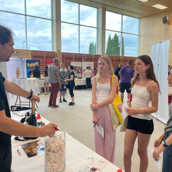 Eine Indoor-Veranstaltung mit einer Glaswand und Bäumen draußen. Ein Mann spricht mit zwei Frauen, während ein Glas mit Süßigkeiten auf einem Tisch vor ihm steht. Mehrere Leute sind unterwegs.