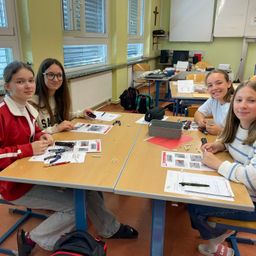 Vier Mädchen sitzen um einen Tisch in einem Klassenzimmer und arbeiten an einem Projekt. Sie haben Papiere und Werkzeuge auf dem Tisch. Das Klassenzimmer hat gelbe Wände und blaue Stühle.