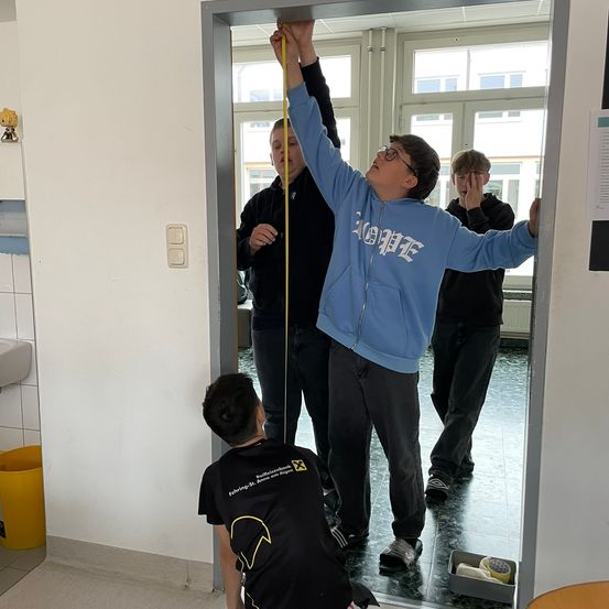Ein Junge misst die Höhe eines Türrahmens, während andere zusehen. Ein anderer Junge kniet und beobachtet. Es gibt ein Waschbecken, einen Mülleimer und einen Schalter an der Wand.