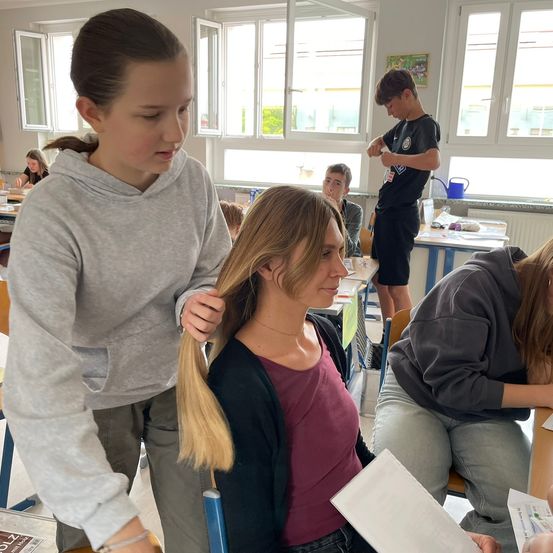Eine Frau mit blonden Haaren lässt sich von einem jungen Mädchen in einem Klassenzimmer die Haare flechten. Andere Schüler sitzen an ihren Schreibtischen, einer steht und hält eine Gießkanne.