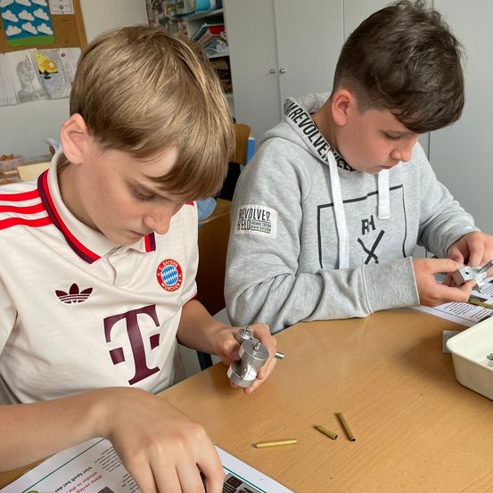 Zwei junge Jungen arbeiten in einem Klassenzimmer an einem Projekt. Ein Junge trägt eine graue Kapuzenjacke mit einem Grafik auf der Vorderseite, und der andere trägt ein weißes Shirt mit einem rot-weiß gestreiften Kragen. Sie halten beide Werkzeuge und scheinen etwas zu reparieren.