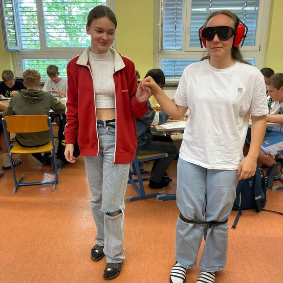 Zwei Mädchen stehen in einem Klassenzimmer, eines in einer roten Jacke und Kopfhörern, das andere in einem weißen T-Shirt mit Logo. Sie halten Händchen, und Schüler sitzen an Tischen im Hintergrund.