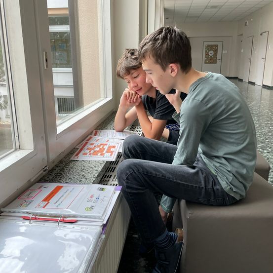 Zwei Jungen sitzen auf einer Bank und schauen sich ein Puzzle auf einem Schreibtisch an. Der Junge auf der rechten Seite benutzt sein Handy. Der Junge auf der linken Seite schaut sich das Puzzle an.