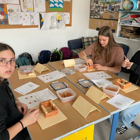 Drei Schüler sitzen an einem Tisch und arbeiten an Ziegelstrukturen. Eines Mädchen baut, während ein anderes auf einem Blatt Papier schreibt. Sie haben mehrere Ziegelstrukturen auf dem Tisch.