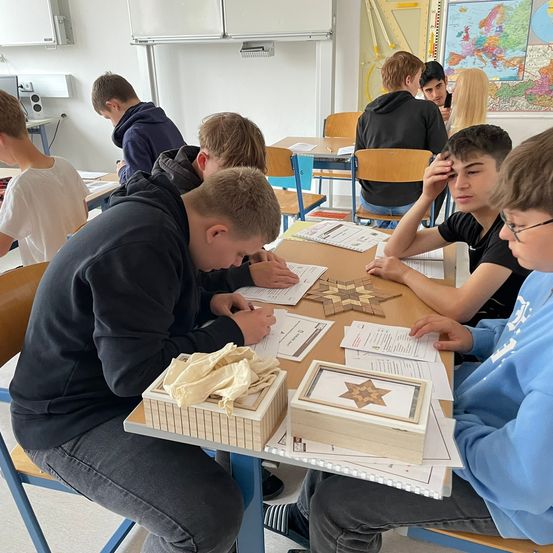 Eine Gruppe von Schülern in einem Klassenzimmer ist mit einer Puzzleaktivität beschäftigt. Ein Schüler beugt sich über einen Tisch mit verschiedenen Puzzleteilen.
