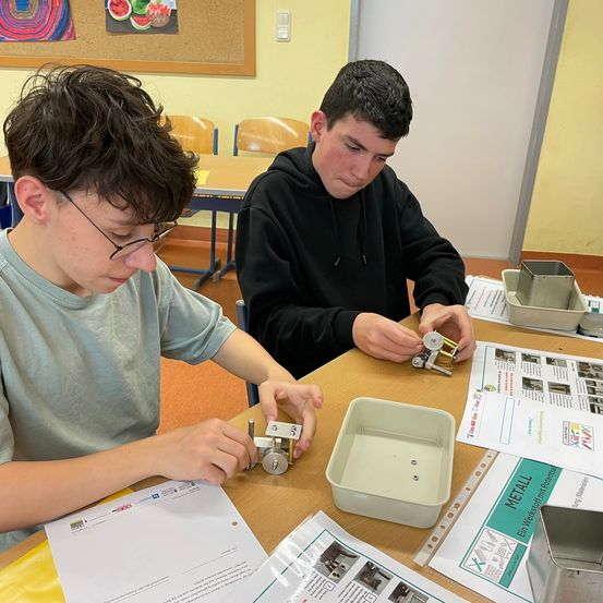 Zwei Schüler arbeiten an einem Projekt an einem Tisch. Ein Schüler trägt eine Brille und konzentriert sich auf ein mechanisches Gerät. Der andere Schüler in einer schwarzen Kapuzenjacke hält ein Metallobjekt. Der Tisch ist mit Papieren, einer weißen Box und einem silbernen Tablett übersät. Eine Pinnwand mit Obstbildern hängt an der Wand.