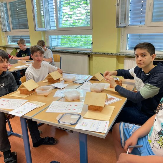 Schüler in einem Klassenzimmer arbeiten an einem Projekt. Sie bauen kleine Häuser aus Ziegelsteinen. Papiere und Plastikbehälter sind auf dem Tisch. Ein Schüler sitzt rechts, ein anderer links.