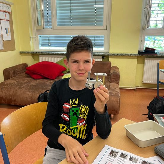 Ein Junge in einem schwarzen Shirt mit einem Minecraft-Design hält ein Metallobjekt. Er sitzt an einem Tisch mit Papieren und einer Schale. Dahinter ist ein Sofa mit Kissen und Fenstern mit Jalousien.