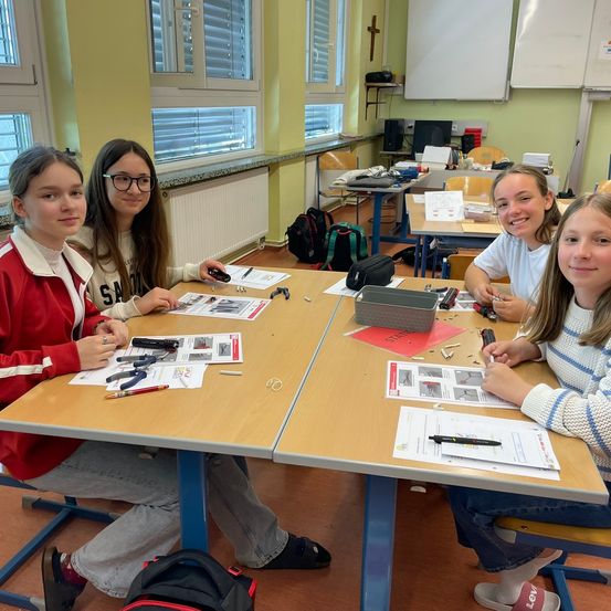 Vier Mädchen sitzen in einem Klassenzimmer an einem Schreibtisch, lächeln und arbeiten an einem Projekt. Papiere, Werkzeuge und eine schwarze Tasche sind auf dem Tisch. Dahinter steht ein weiterer Schreibtisch mit einem Rucksack und einer Tafel.