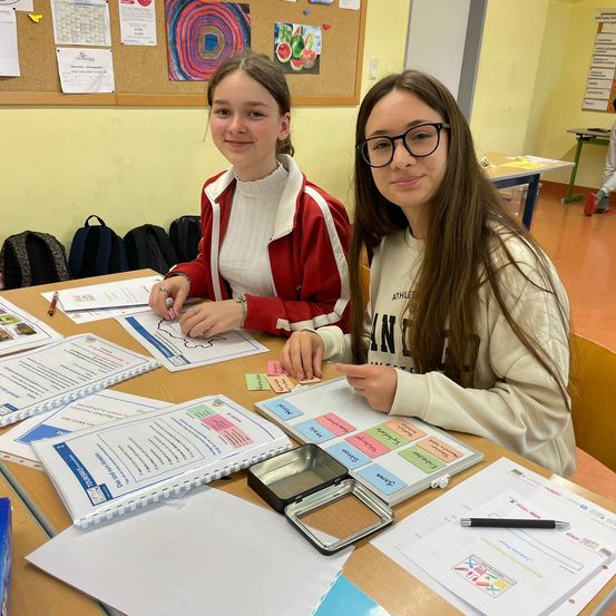 Zwei Mädchen sitzen lächelnd an einem Schreibtisch, umgeben von Papieren, Haftnotizen und einem Notizbuch. Der Tisch ist mit Materialien für ein Projekt übersät. Ein Bulletin-Board ist hinter ihnen sichtbar.