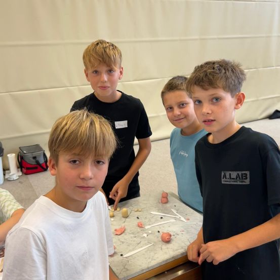 Vier Jungen stehen hinter einem Tisch in einem Klassenzimmer. Ein Junge berührt Gegenstände auf dem Tisch, während die anderen zuschauen.