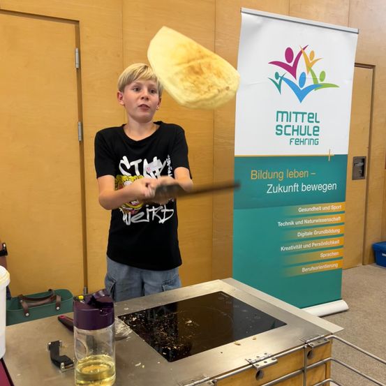 Ein Junge flippt eine Tortilla bei einem Kochwettbewerb. Er steht vor einem Banner mit dem Text 'Mittel Schule Fehringen'. Es gibt einen Tisch mit verschiedenen Gegenständen darauf.