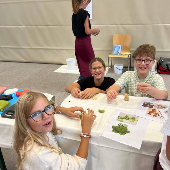 Drei Kinder sitzen an einem Tisch und arbeiten an einem Bastelprojekt mit Ton und Papier. Ein Kind hält eine Froschfigur. Eine Frau steht hinter ihnen.