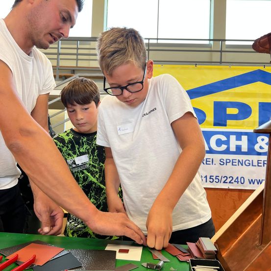 Drei junge Männer sind in einer Werkstatt um einen Tisch versammelt. Zwei Jungen tragen weiße T-Shirts, einer davon mit einer Brille. Der Junge auf der linken Seite trägt ein grünes Shirt. Sie arbeiten an einem Projekt mit verschiedenen Werkzeugen und Materialien auf dem Tisch. Ein Banner mit dem Text 'Spenglerei Spenglerei' ist im Hintergrund zu sehen.