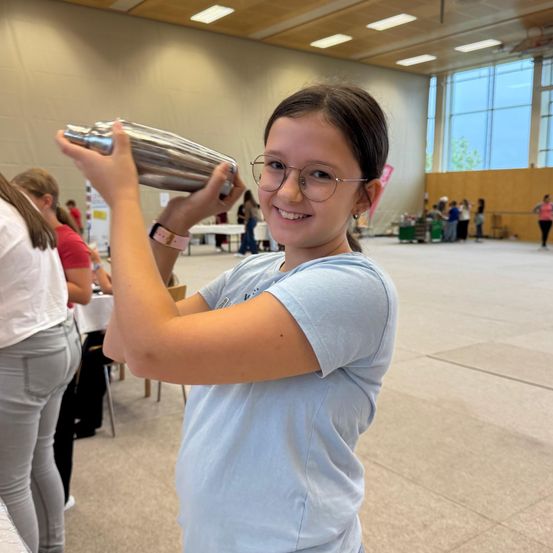 Ein Mädchen mit Brille lächelt und hält einen silbernen Shaker in einer Halle. Hinter ihr sind Tische, Stühle und Menschen sowie große Fenster mit Bäumen draußen.