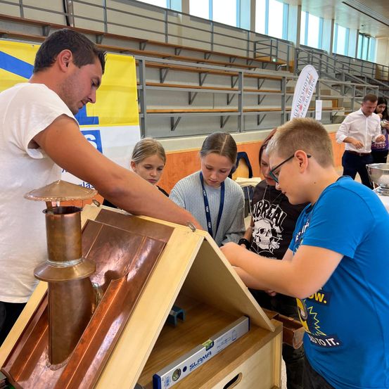 Ein Mann demonstriert einen Kupferschornstein an einem Holzmodellhaus für eine Gruppe von Schülern in einer Innenumgebung.