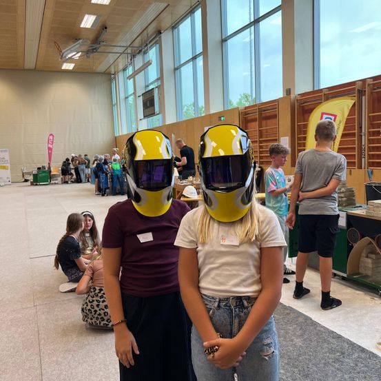 Zwei Mädchen mit gelben Helmen stehen in einem großen Saal mit mehreren Leuten um sie herum. Ein Junge steht mit verschränkten Armen hinter ihnen.