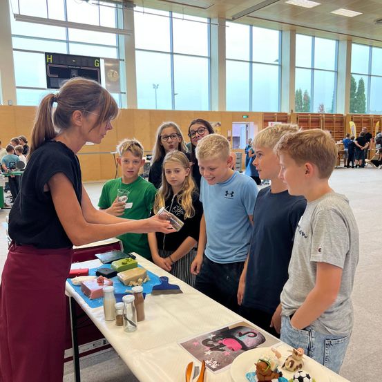 Eine Frau in einem schwarzen Shirt und einer roten Hose demonstriert etwas für eine Gruppe von Kindern, die um einen Tisch mit verschiedenen Gegenständen darauf in einer großen Halle stehen.