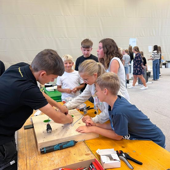 Mehrere Kinder arbeiten in einem Klassenzimmer mit Werkzeugen an einem Holzbrett. Ein Junge in Schwarz bohrt, während ein anderer Junge in Blau zusieht. Ein Mädchen in einem weißen T-Shirt beobachtet. Dahinter stehen noch einige weitere Kinder.