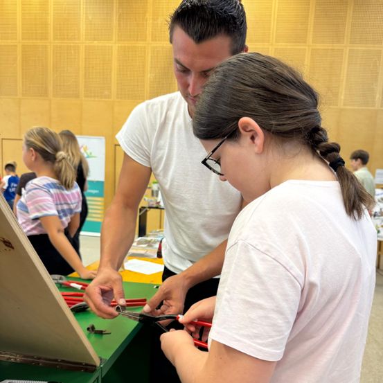 Ein Mann und ein Mädchen benutzen eine Schere auf einem grünen Tisch. Andere Leute sind um sie herum.