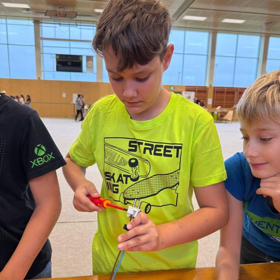 Drei Jungen arbeiten drinnen an einem Projekt. Der mittlere Junge hält einen Stecker und einen Schraubendreher. Der Junge links trägt ein schwarzes Shirt mit einem Xbox-Logo. Der Junge rechts trägt ein blaues Shirt.