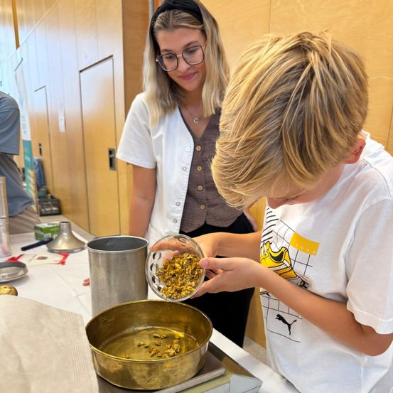 Ein junger Junge hält eine Schüssel mit gelben Blumen, während eine Frau ihn in einem Raum mit Holzwänden beobachtet.