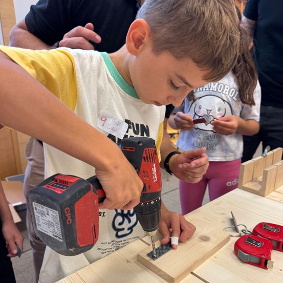 Ein Junge benutzt eine Bohrmaschine, um an einem Holzprojekt zu arbeiten, trägt ein Shirt mit dem Wort 'FUN'. Eine Mädchen in der Nähe hält ein kleines Objekt, möglicherweise ein Bastelmaterial.