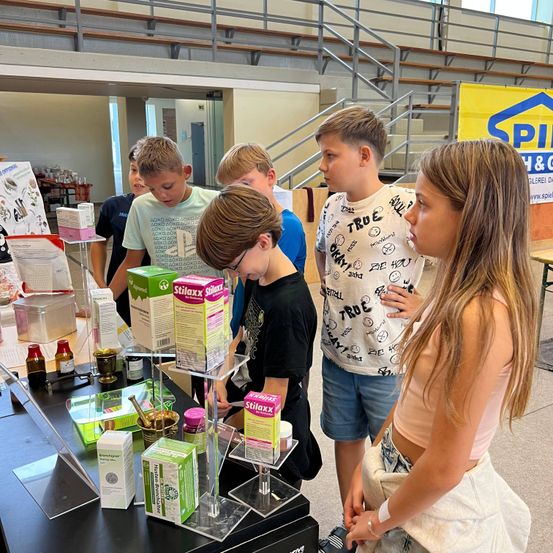 Eine Gruppe von Kindern schaut sich Produkte auf einem Tisch bei einer Veranstaltung an. Sie stehen um eine Ausstellung mit verschiedenen Artikeln und einem Banner.