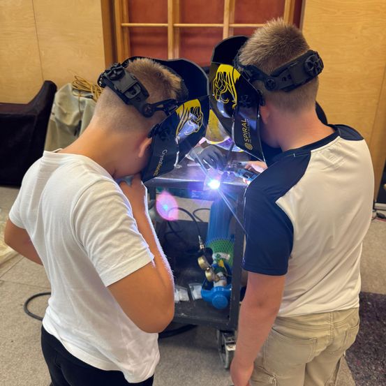 Zwei junge Jungen, die Schweißmasken tragen, sind mit einer Schweißaktivität beschäftigt. Ein Junge beobachtet, während der andere an der Schweißmaschine arbeitet. Sie stehen in einem Raum mit einer Holzwand und einer Couch im Hintergrund.