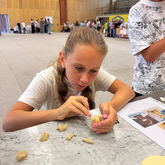 Ein junges Mädchen sitzt an einem Tisch und formt konzentriert Teig zu kleinen, runden Formen. Hinter ihr ist ein geschäftiger Raum zu sehen, mit Menschen und Ständen im Hintergrund.