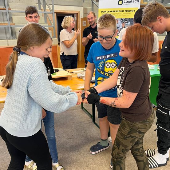Mehrere Teenager interagieren in einem Raum mit einem Tisch. Ein Mädchen mit Brille schüttelt die Hand eines Jungen im blauen Hemd. Eine Frau hinter ihnen klatscht. Ein Werbeschild ist im Hintergrund.