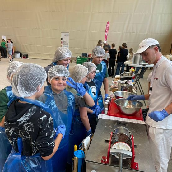 Mehrere Personen in Haarnetzen und Schürzen stehen um einen Tisch, an dem ein Mann in einem weißen Hut und Handschuhen etwas in einer Schüssel mischt.
