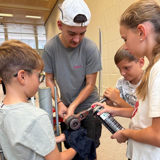 Drei junge Personen arbeiten gemeinsam an einem Projekt in einem Gebäude. Ein Junge hält eine Sprühdose, während ein anderer ein mechanisches Werkzeug hält. Ein Mädchen in der Mitte beobachtet aufmerksam.