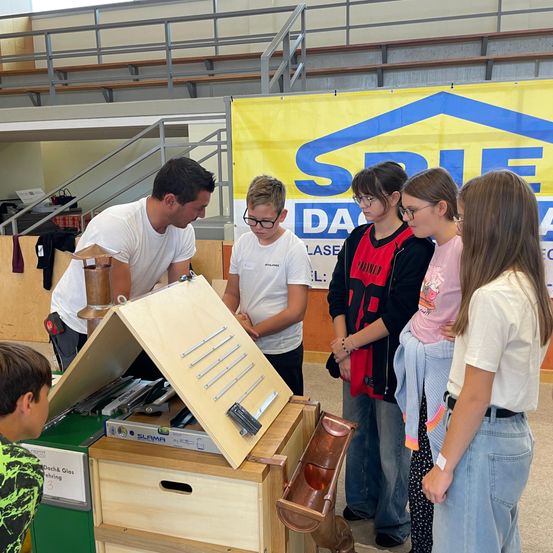Ein Mann und mehrere Kinder untersuchen eine Holzstruktur mit Kupferdach bei einer Veranstaltung im Innenbereich, begleitet von einem großen Banner mit Text.