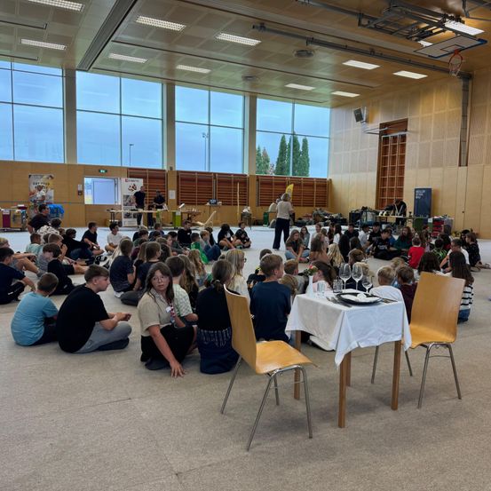 Eine große Gruppe von Kindern sitzt auf dem Boden in einem Raum mit großen Fenstern. Eine Frau steht in der Mitte des Raumes, möglicherweise gibt sie eine Präsentation. Ein Tisch mit Weingläsern und Tellern ist vor den Kindern aufgebaut.