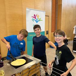 Drei Jungen kochen drinnen ein Omelett auf einem kleinen Induktionsherd. Der Junge auf der linken Seite wende das Omelett. Der Junge in der Mitte schaut zu. Der Junge auf der rechten Seite lächelt. Ein Banner steht hinter ihnen.