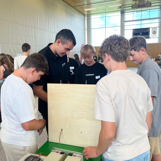 Fünf junge Jungen in weißen Hemden schauen auf eine Holzplatte mit einem Mann in einem schwarzen Hemd. Sie sind in einem Raum mit einer weißen Wand und Glasfenstern.