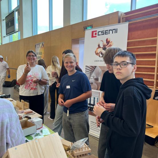 Eine Gruppe junger Menschen steht um einen Tisch mit Holzmodellen. Ein Banner mit dem Wort 'CSERNI' steht dahinter. Eine Frau in einem weißen T-Shirt hält ein Holzmodell.