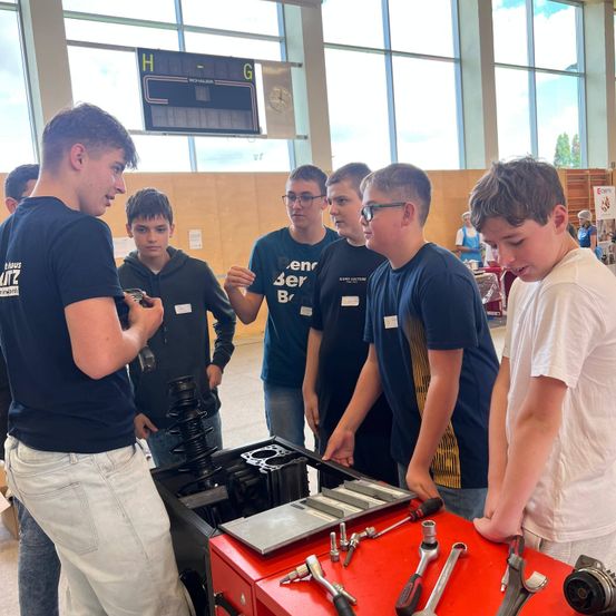 Eine Gruppe junger Männer in einem Raum, wahrscheinlich einem Klassenzimmer, stehen um eine rote Werkzeugkiste herum. Sie betrachten die Werkzeugkiste und einige von ihnen halten Werkzeuge. Der Mann in der Mitte hält ein Werkzeug in der Hand. Der Raum hat eine Holzwand und eine Glaswand mit Blick nach draußen.