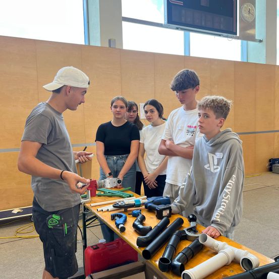 Eine Gruppe junger Leute steht um einen Tisch mit verschiedenen Werkzeugen. Der Mann in der weißen Kappe erklärt dem Jungen in der grauen Kapuzenjacke etwas.