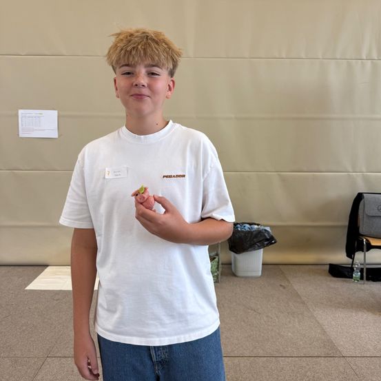 Ein junger Junge mit blonden Haaren steht drinnen und hält eine rosa Figur. Er trägt ein weißes T-Shirt mit Logo und blaue Jeans. Hinter ihm ist eine beige Wand mit einem Papier und einem Mülleimer.