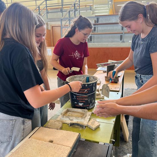 Vier Mädchen arbeiten gemeinsam an einem Bauprojekt. Eine mischt Zement, während die anderen Materialien vorbereiten. Sie befinden sich in einem Raum mit Holzboden und Tribünen im Hintergrund.