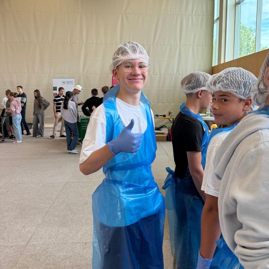 Ein junger Mann in einem blauen Schutzanzug gibt ein Daumen hoch, während andere in ähnlichen Anzügen in einem großen Raum zusehen.