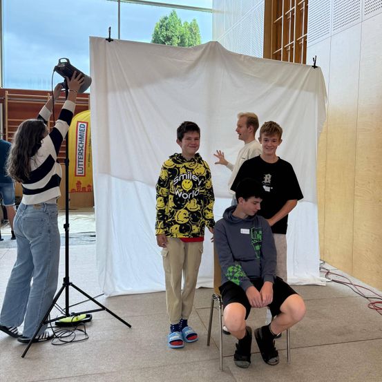 Eine Frau hält eine Kamera, während vier Jungen vor einem weißen Hintergrund stehen und sitzen, wahrscheinlich für ein Fotoshooting. Bäume sind durch ein Glasfenster sichtbar.