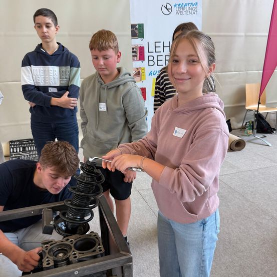 Eine Gruppe von Schülern, darunter ein Mädchen in einer rosa Kapuzenjacke, arbeiten an einem mechanischen Projekt. Ein Junge benutzt einen Schraubenschlüssel an einer Metallstruktur.