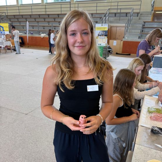 Ein junges Mädchen mit blonden Haaren lächelt in einem Innenraum, während es ein kleines rosa Objekt in der Hand hält. Hinter ihr sind andere Kinder und Erwachsene, die sich mit Aktivitäten beschäftigen.