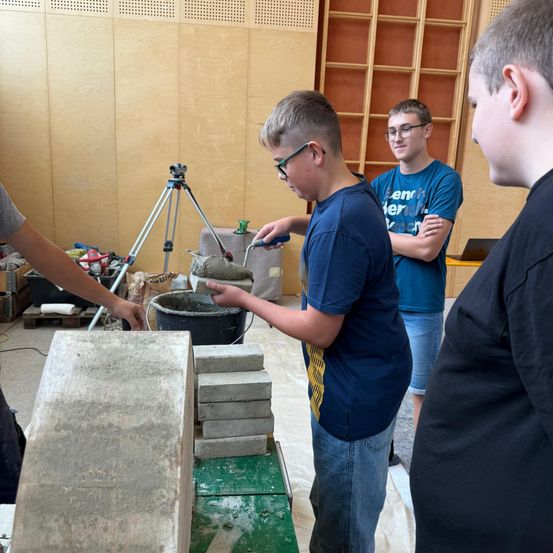 Drei Jungen arbeiten an einem Zementblockprojekt. Ein Junge benutzt eine Kelle, um Zement zwischen die Blöcke zu auftragen. Ein anderer Junge beobachtet, während der dritte Junge zusieht.