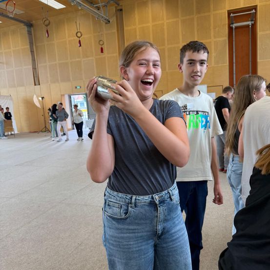 Ein Mädchen trinkt aus einem silbernen Becher und lacht, während es in einer Turnhalle steht, mit anderen Schülern im Hintergrund.