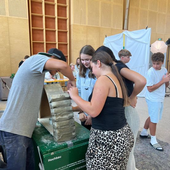 Eine Gruppe junger Menschen arbeitet gemeinsam an einer grünen Box, wahrscheinlich in einem schulischen Umfeld. Der Junge in Grau schaut auf die Box, während das Mädchen im Leopardenmuster zusieht.